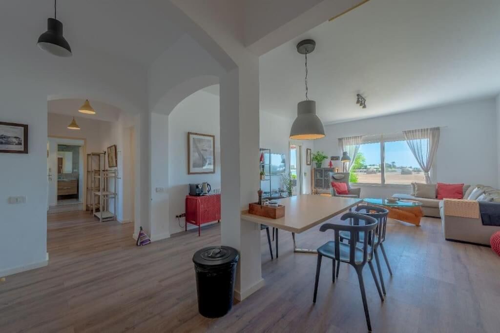 A spacious living area with a dining table, chairs, and a sofa. There is a kitchen area in the background with a red cabinet and a hanging light fixture.