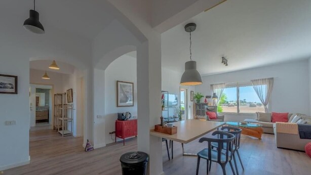 A spacious living area with a dining table, chairs, and a sofa. There is a kitchen area in the background with a red cabinet and a hanging light fixture. - unique flat_ garden and lagoon (Hurghada)