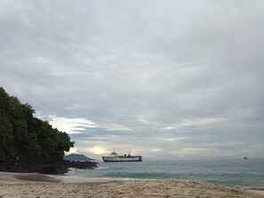 Une plage avec un bateau visible au loin, une côte rocheuse et un ciel nuageux.