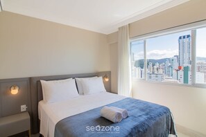 A hotel room with a bed, a nightstand, a window with a city view, and a wall-mounted light. - Apart w/ Side Sea View in Front of Beach Iptr0241 (Balneário Camboriú)