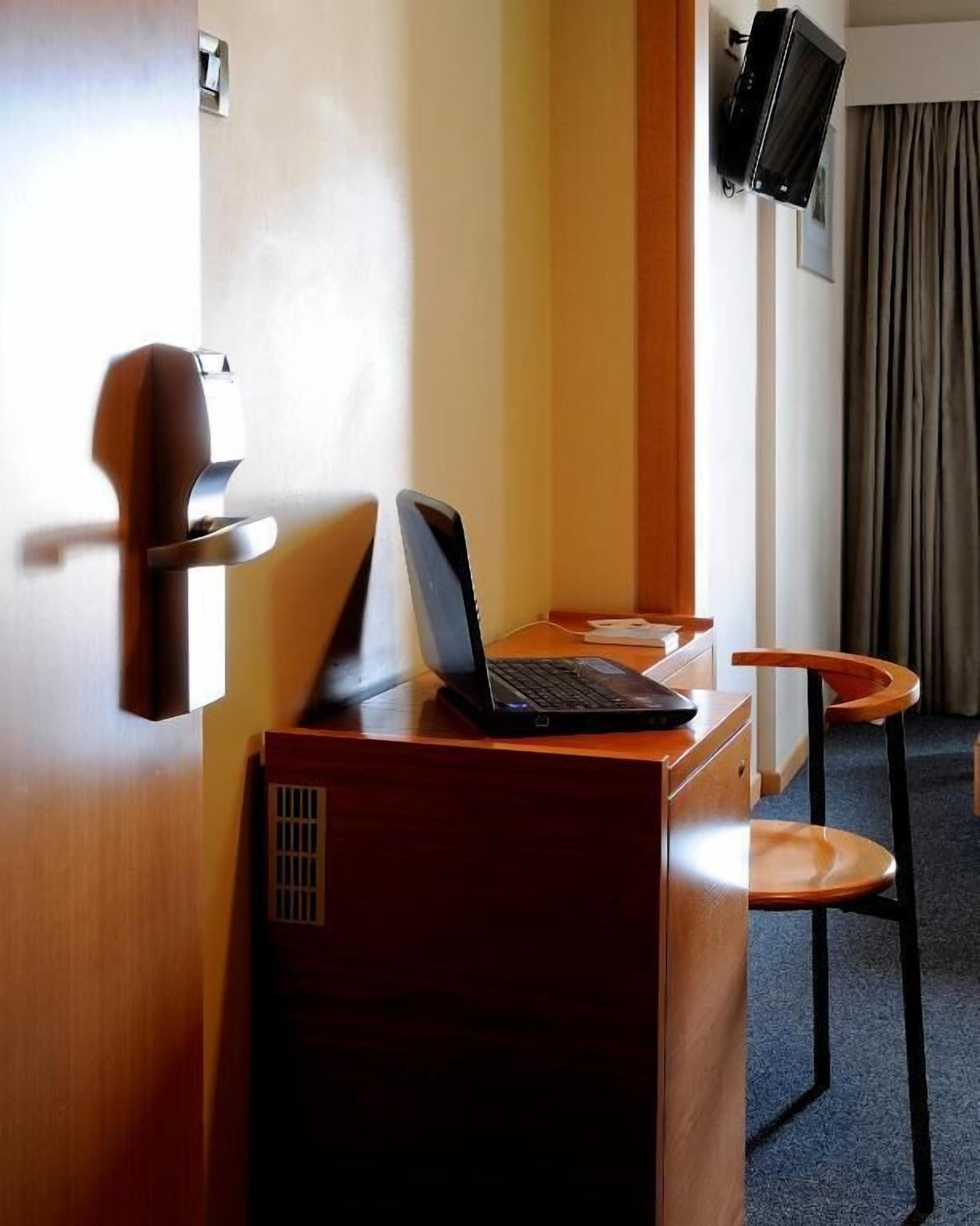 A hotel room with a desk, a laptop, a chair, and a television mounted on the wall.