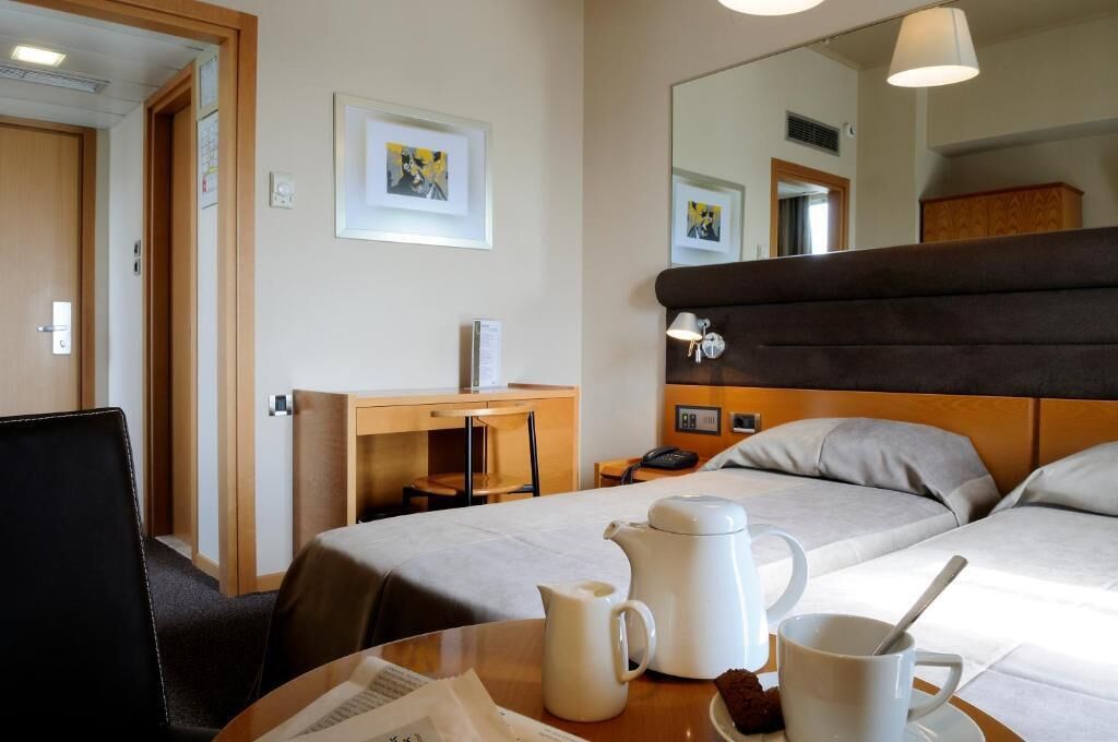 A hotel room with a bed, a desk, a chair, a table with a coffee pot and mug, and a mirror.