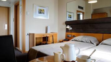 A hotel room with a bed, a desk, a chair, a table with a coffee pot and mug, and a mirror.