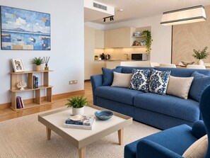 A modern living room with a blue sofa, a coffee table with books and a bowl, a bookshelf, and a painting on the wall. - Mare Blu - Portopiccolo Apartments (Sistiana)