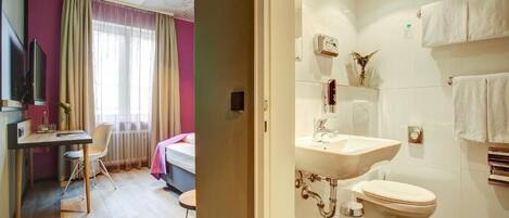 A hotel room with a bed, a desk, a chair, a window with curtains, and a bathroom with a sink, toilet, and towel rack.