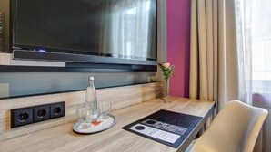 A hotel room with a flat-screen TV mounted on the wall, a wooden desk with a tray table, a vase with a single flower, and a glass of water.