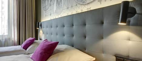A hotel room with a tufted headboard, two beds with pillows, a wall-mounted light fixture, and a geometric patterned wall decoration.
