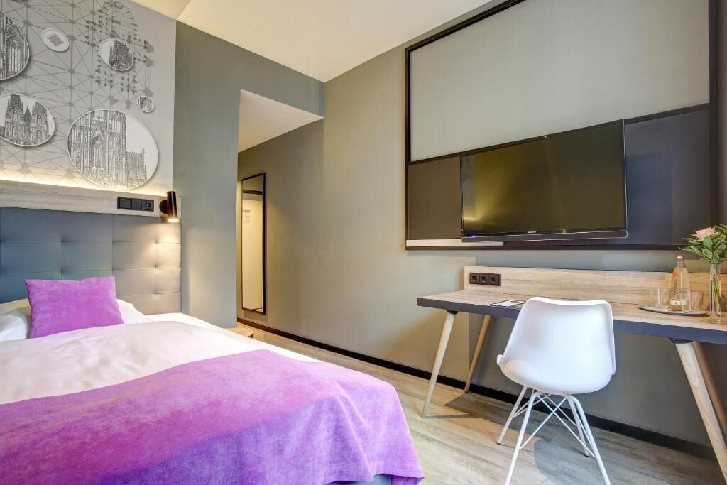 Habitación de hotel moderna con cama, escritorio, silla, televisión de pantalla plana y un cuadro circular en la pared.