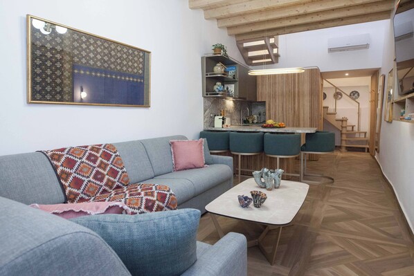 A modern living room with a grey sofa, a coffee table, and a kitchen area with bar stools. - Exclusive Syntagma & Plaka 2BD Townhouse (Athina)