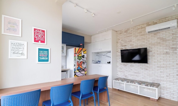 A modern kitchen with a colorful refrigerator, a dining area with blue chairs, and a TV mounted on a brick wall. - Apt Foot in the Sand in Jurere Jbm201 (Florianópolis)