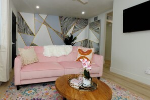 Ein modernes Wohnzimmer mit einem rosafarbenen Sofa, dekorativen Kissen, einem Holztisch mit Büchern und einer Vase, sowie einer Wand mit geometrischem Muster.