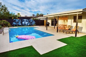 Ein Schwimmbecken mit Wandgemälde, eine überdachte Terrasse mit Gartenmöbeln und ein gepflegter Rasen.