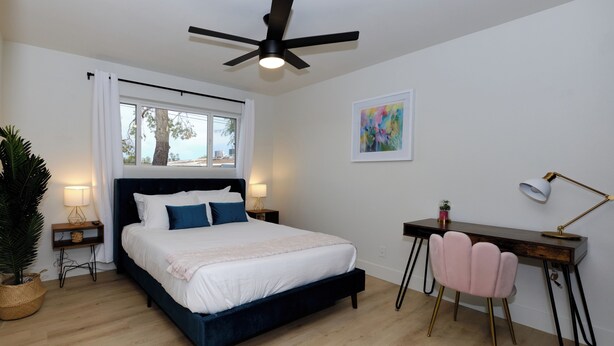 Ein Schlafzimmer mit Bett, Schreibtisch, Stuhl, Deckenventilator, Fenster und Kunstwerken.