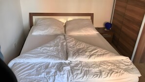 Una cama con ropa blanca, un cabecero de madera y una mesita de noche con un objeto azul.