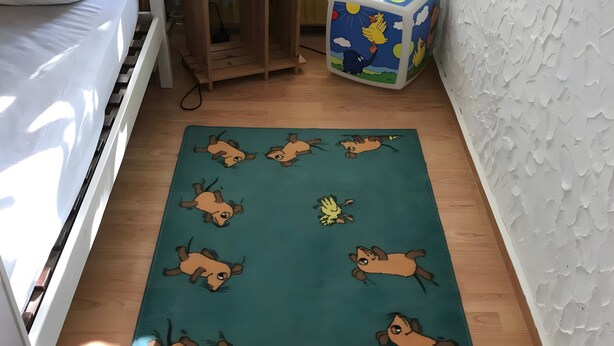 Huoneessa on puulattia, turkoosi, sarjakuvahahmoilla kuvioitu matto, valkoinen seinä, jossa on kuvioitu pinta, ja värikäs kuutionmuotoinen esine.