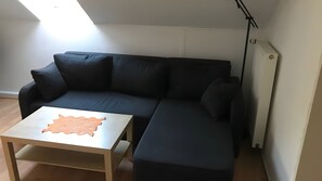 A modern living room with a dark-colored sectional sofa, a small wooden table, and a ceiling light fixture. - Holiday Apartment "Spiekeroog" – 1 Bedroom (Dornum)