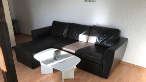 A black leather sectional sofa with a white coffee table in a room with wooden flooring and white textured walls. - Apartment "Baltrum" – 1 Bedroom (Dornum)