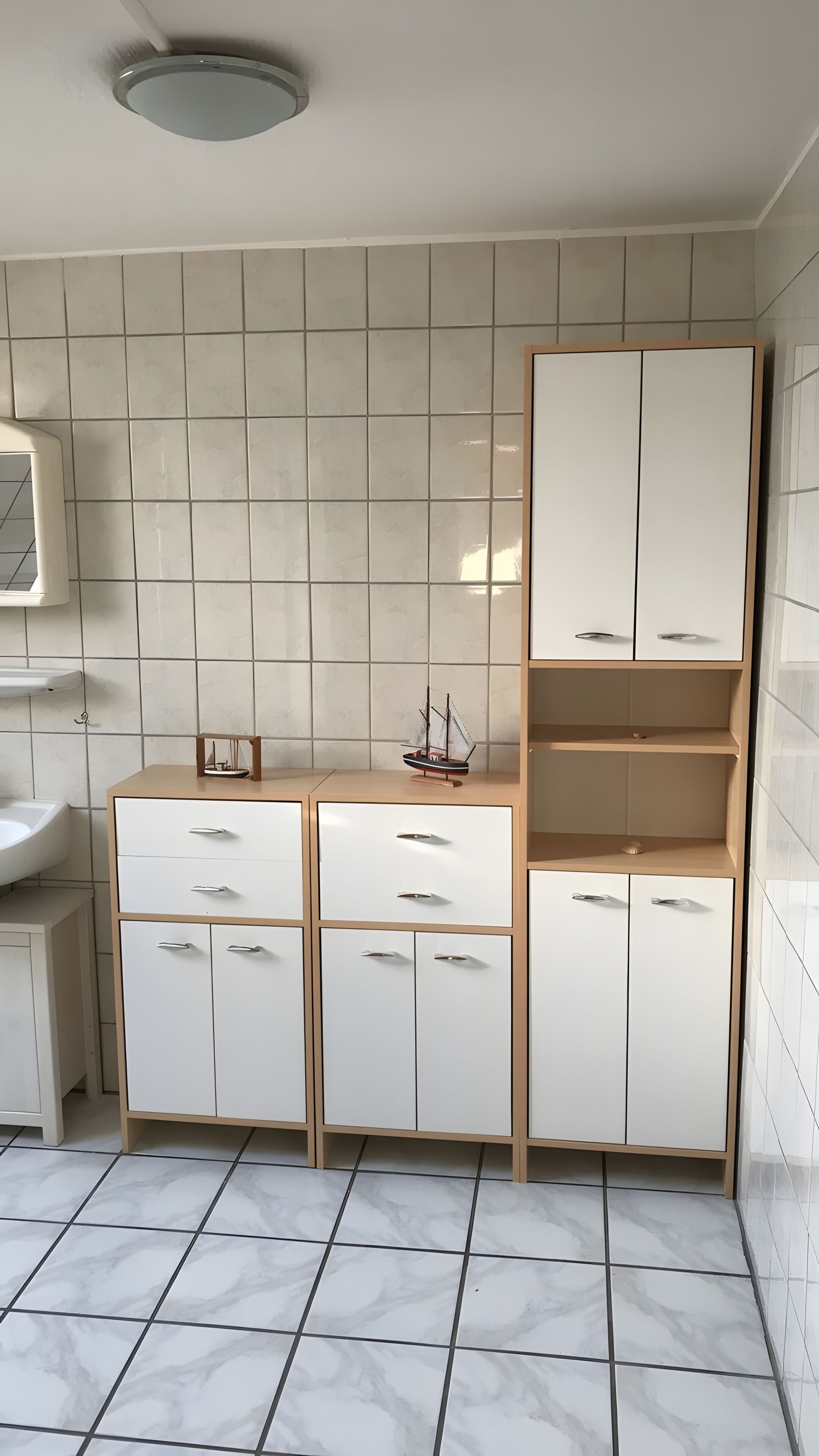 Ein Badezimmer mit weiß geflieste Wand, einem weißen Waschbecken und einem Schrank mit Schubladen und Regalen.