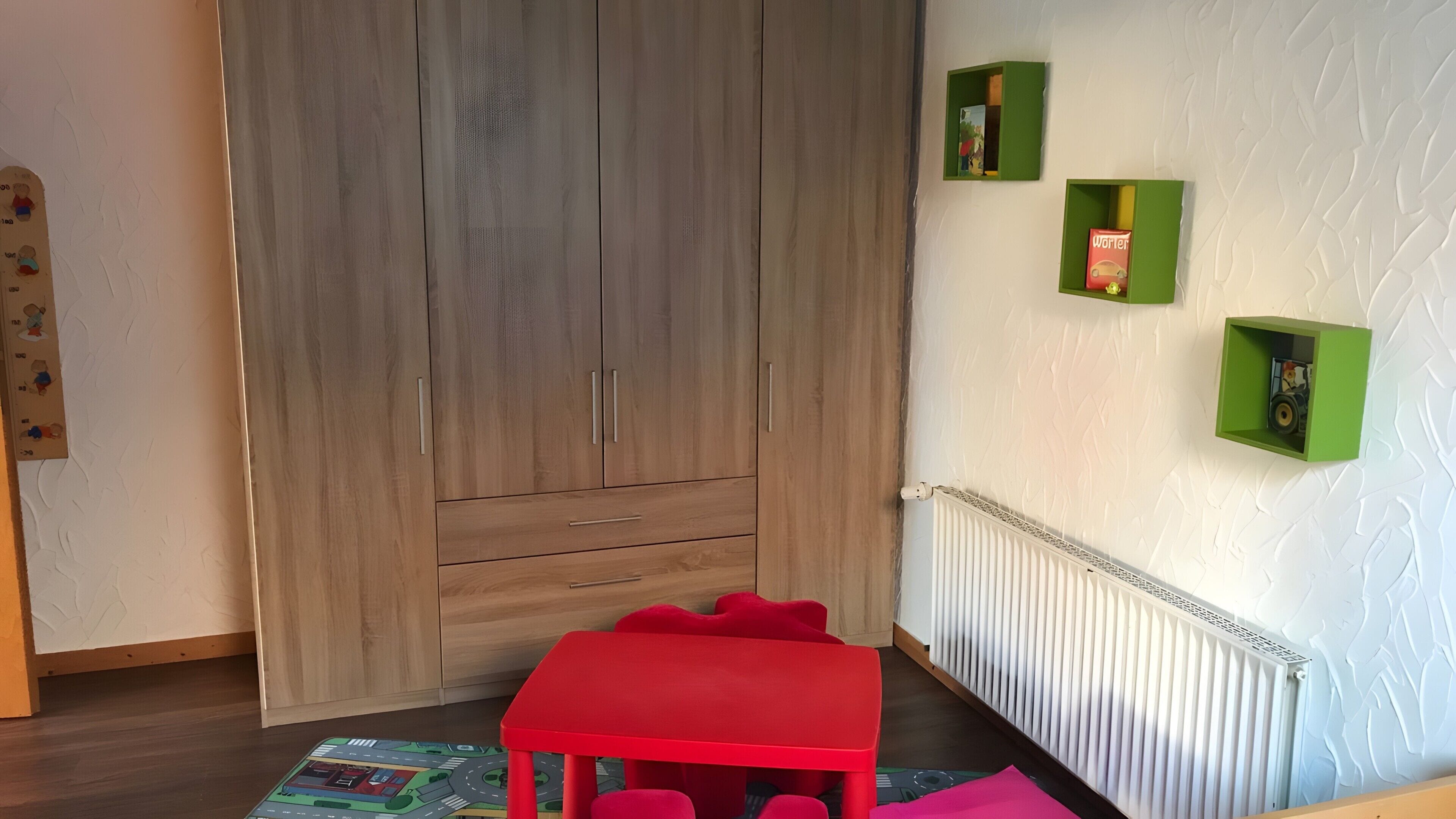 A room with a wooden wardrobe, a red table, and green wall shelves.
