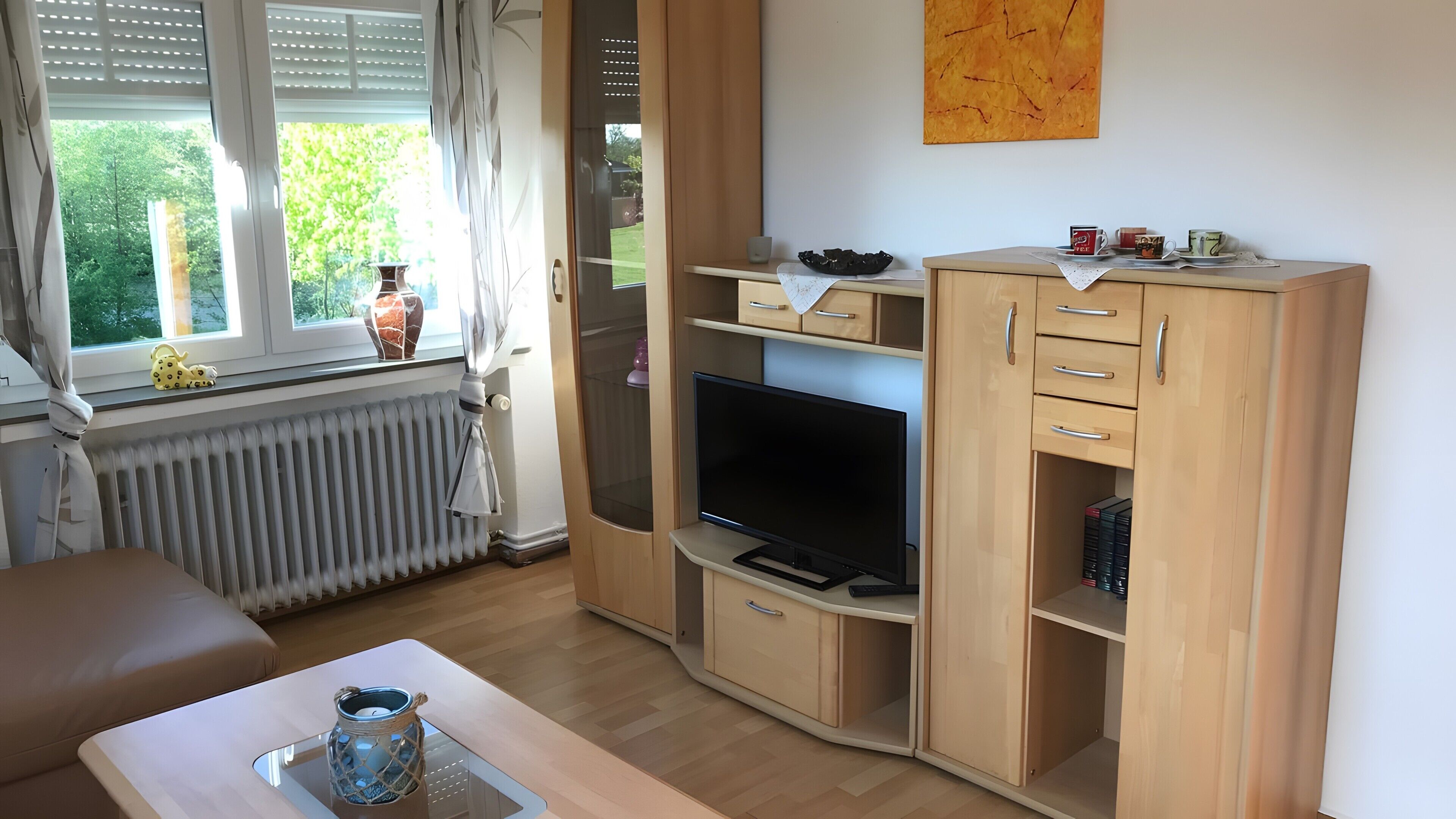 Ein Zimmer mit Fernseher, Holzschränken, Heizkörper und einem mit Vorhängen versehenen Fenster.