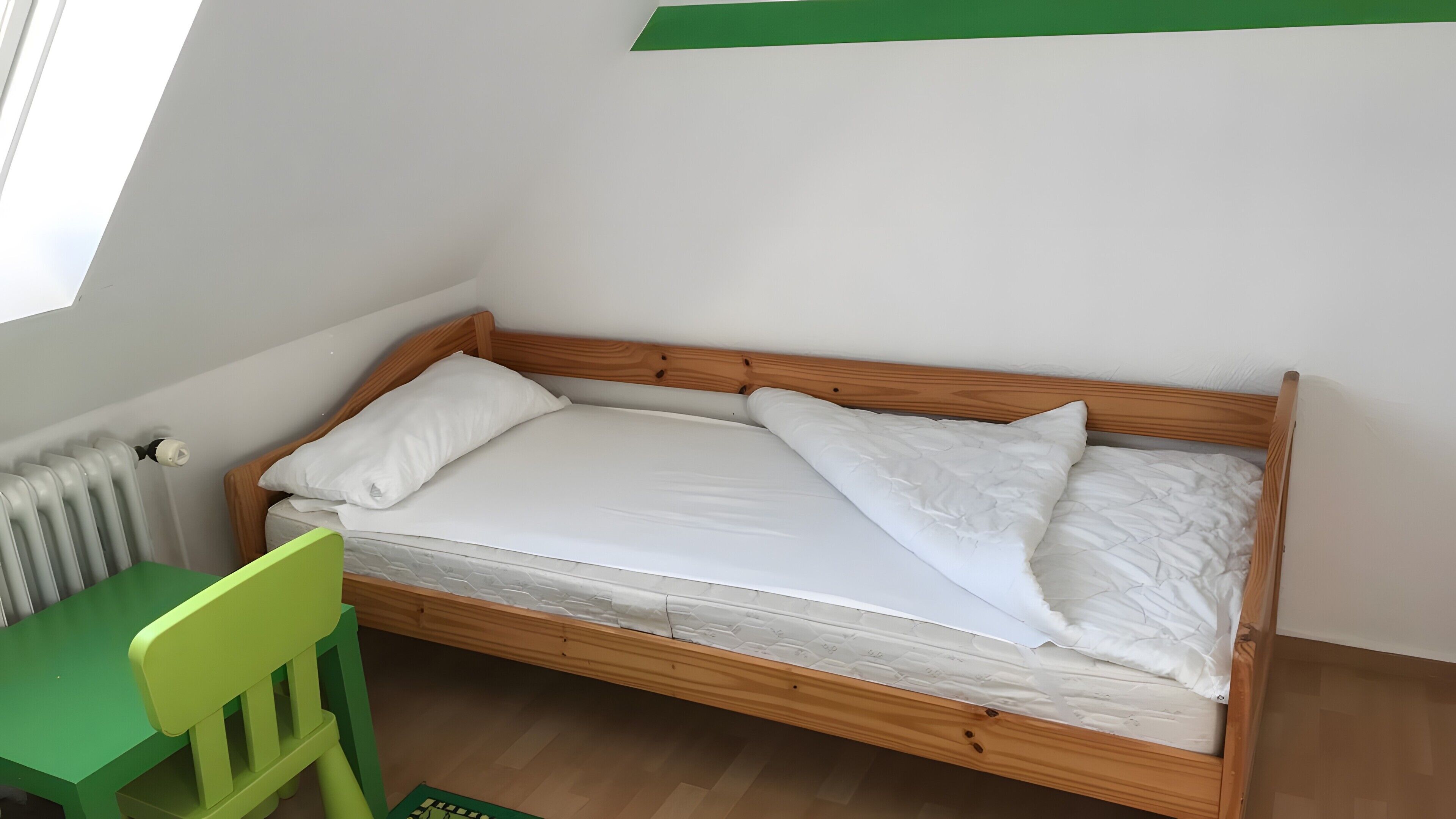A single bed with wooden headboard and white bedding, a green chair, and a radiator on the wall.