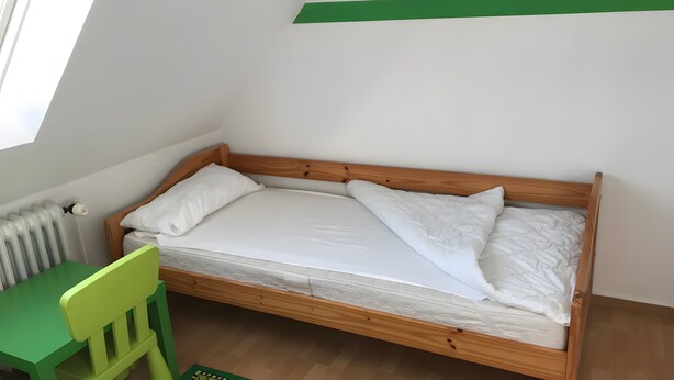 Una cama individual con cabecera de madera y ropa de cama blanca, una silla verde y un radiador en la pared.