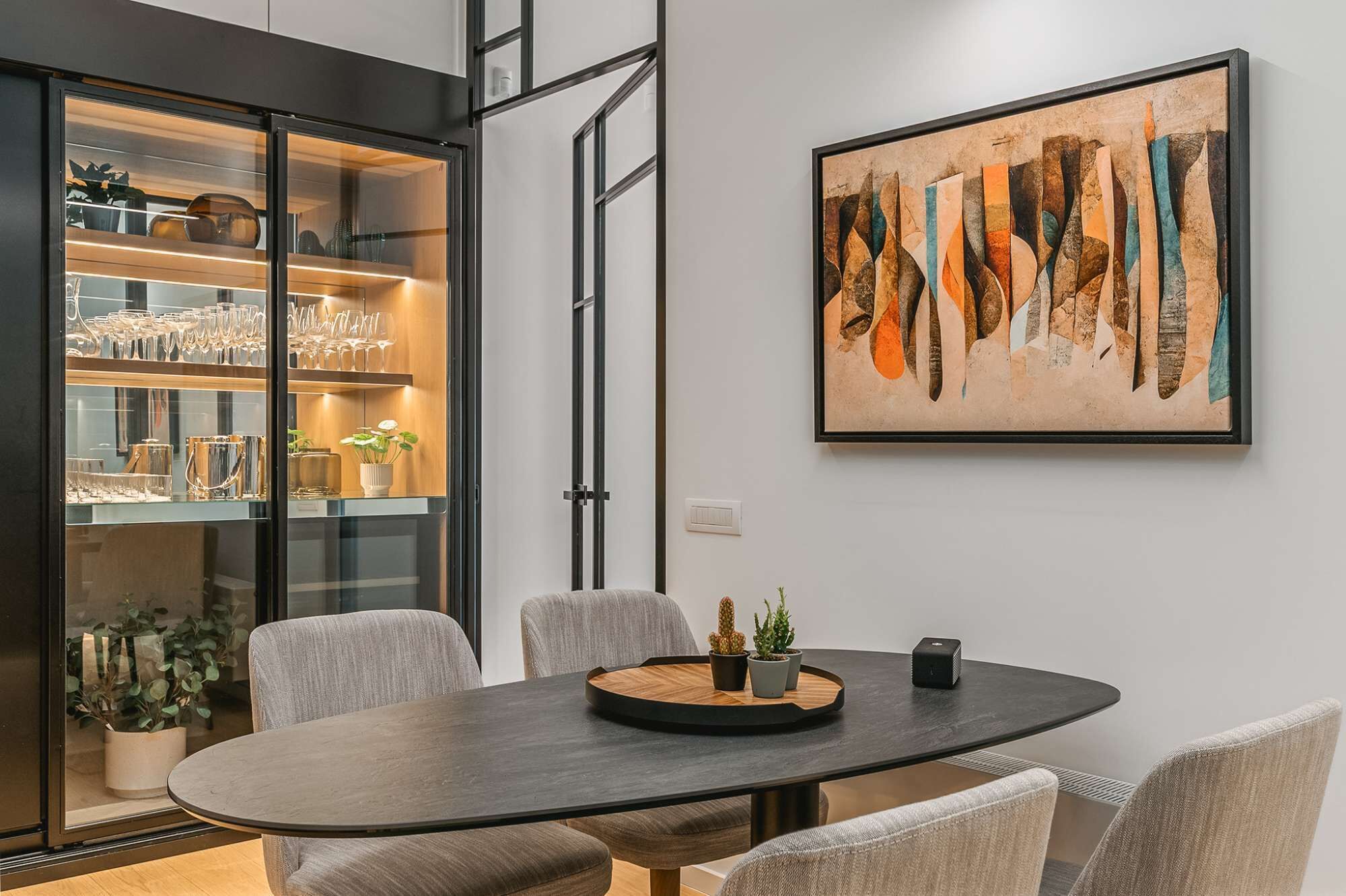 Ein moderner Essbereich mit einem runden Tisch, gepolsterten Stühlen, einer Glasvitrine mit Geschirr und einem gerahmten abstrakten Gemälde an der Wand.