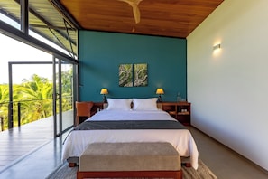 A modern hotel room with a large bed, two bedside lamps, a painting on the wall, and a balcony with a view of palm trees. - BlackRoots Unawatuna (Galle)