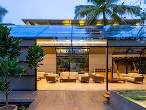 A modern building with a glass facade, outdoor seating area with sofas and tables, and a wooden deck. - BlackRoots Unawatuna (Galle)