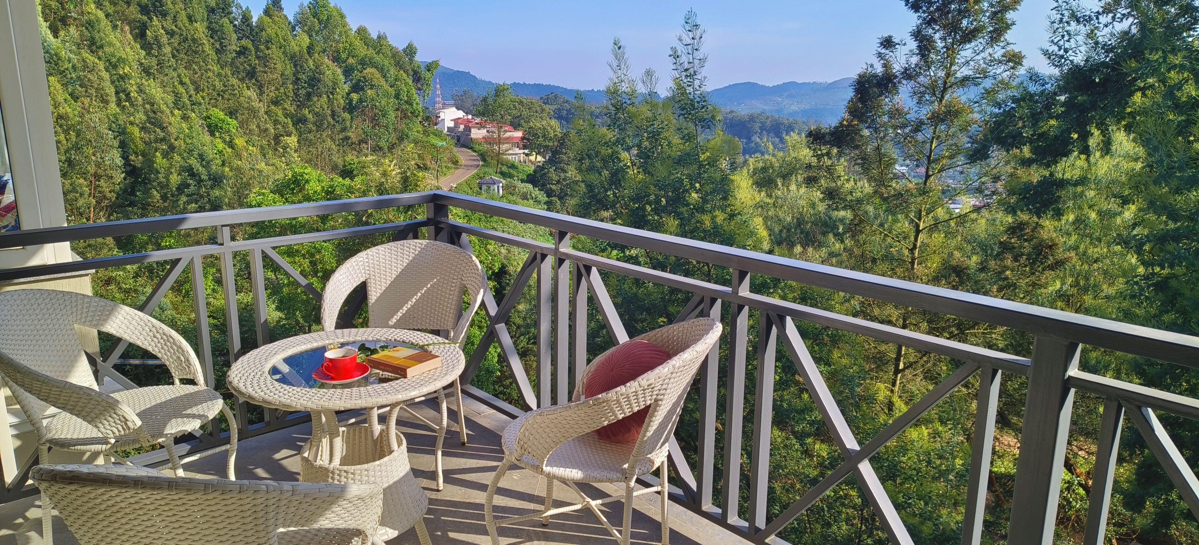 A balcony with wicker furniture and a table, overlooking a forested area with a view of distant buildings.