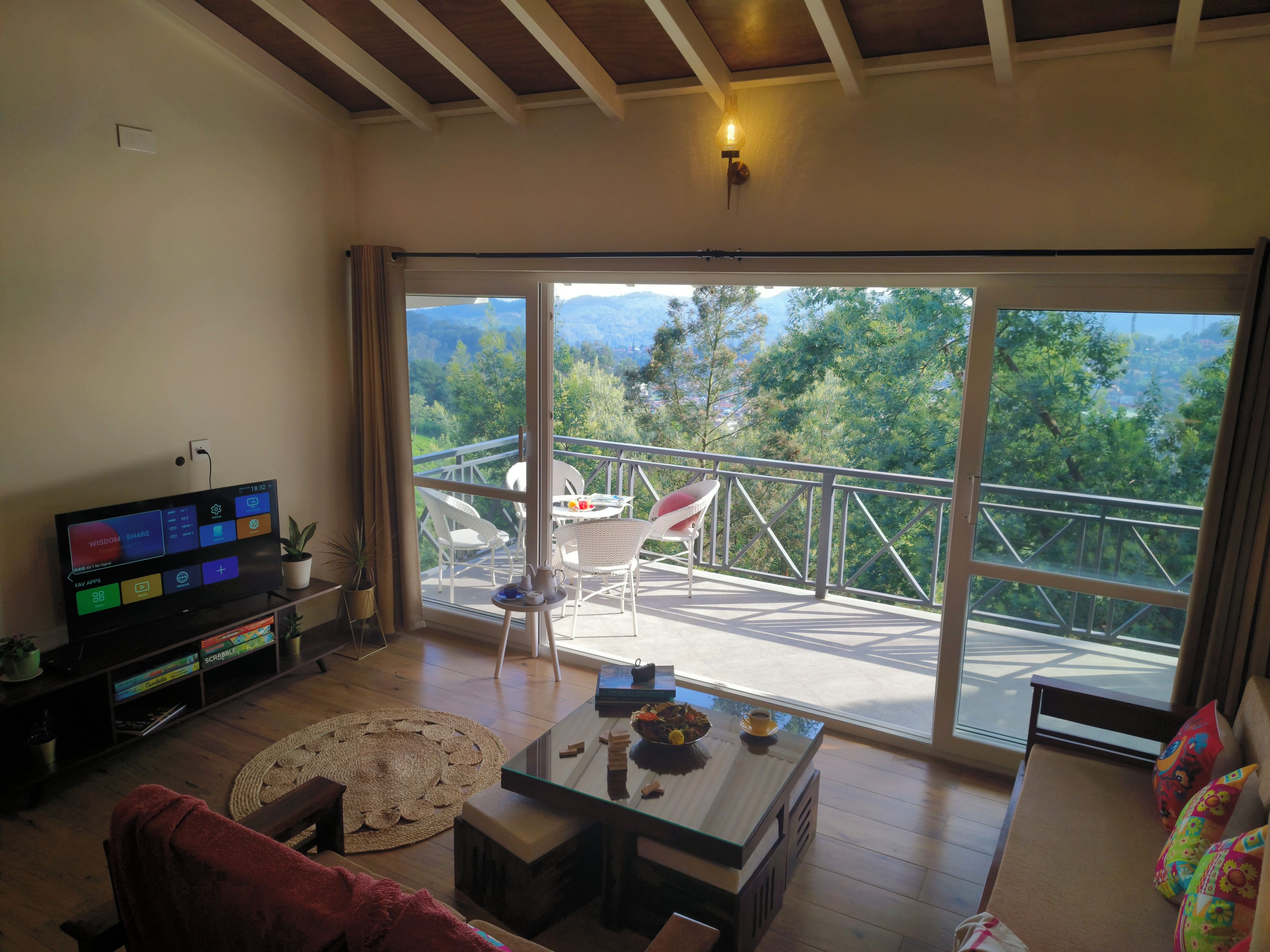 A living room with a television, a glass coffee table, and a sofa. There is a balcony with a view of trees and mountains.