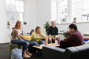 Eine Gruppe von Menschen sitzt in einem modernen, hellen Raum mit großen Fenstern, einem Holztisch und verschiedenen Pflanzen.