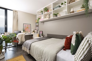 A modern bedroom with a bed, a desk, a chair, and a shelving unit with plants. - Student Only Causeway View (Aberdeen)
