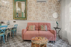 Ein Zimmer mit einem rosafarbenen Sofa, einem Glastisch, einem blauen Sessel, einem runden Spiegel und einem gemusterten Boden.