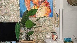 Zona de recepción con una planta en maceta, un escritorio con computadora, un certificado enmarcado y una estantería con libros y un vaso.