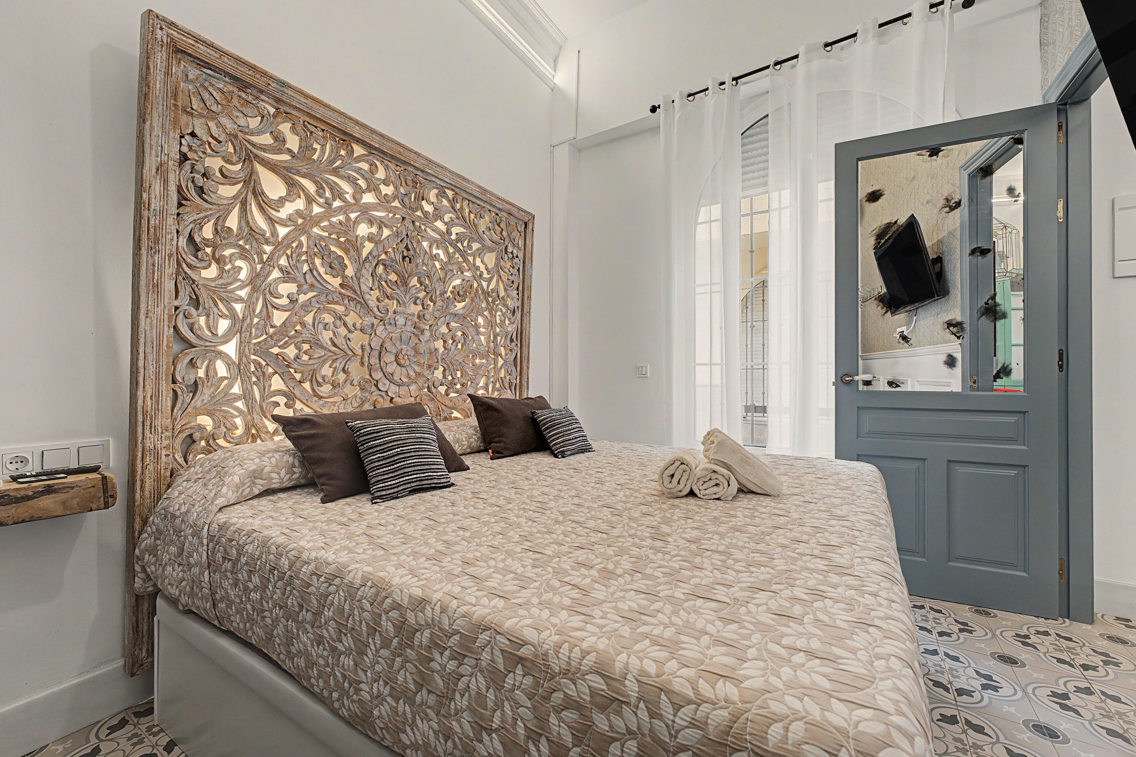 Une chambre à coucher avec une tête de lit décorative, un lit recouvert d’une couvre-lit à motifs, deux oreillers, une table de chevet avec une télécommande, et une porte dotée d’un miroir et d’un porte-manteau.