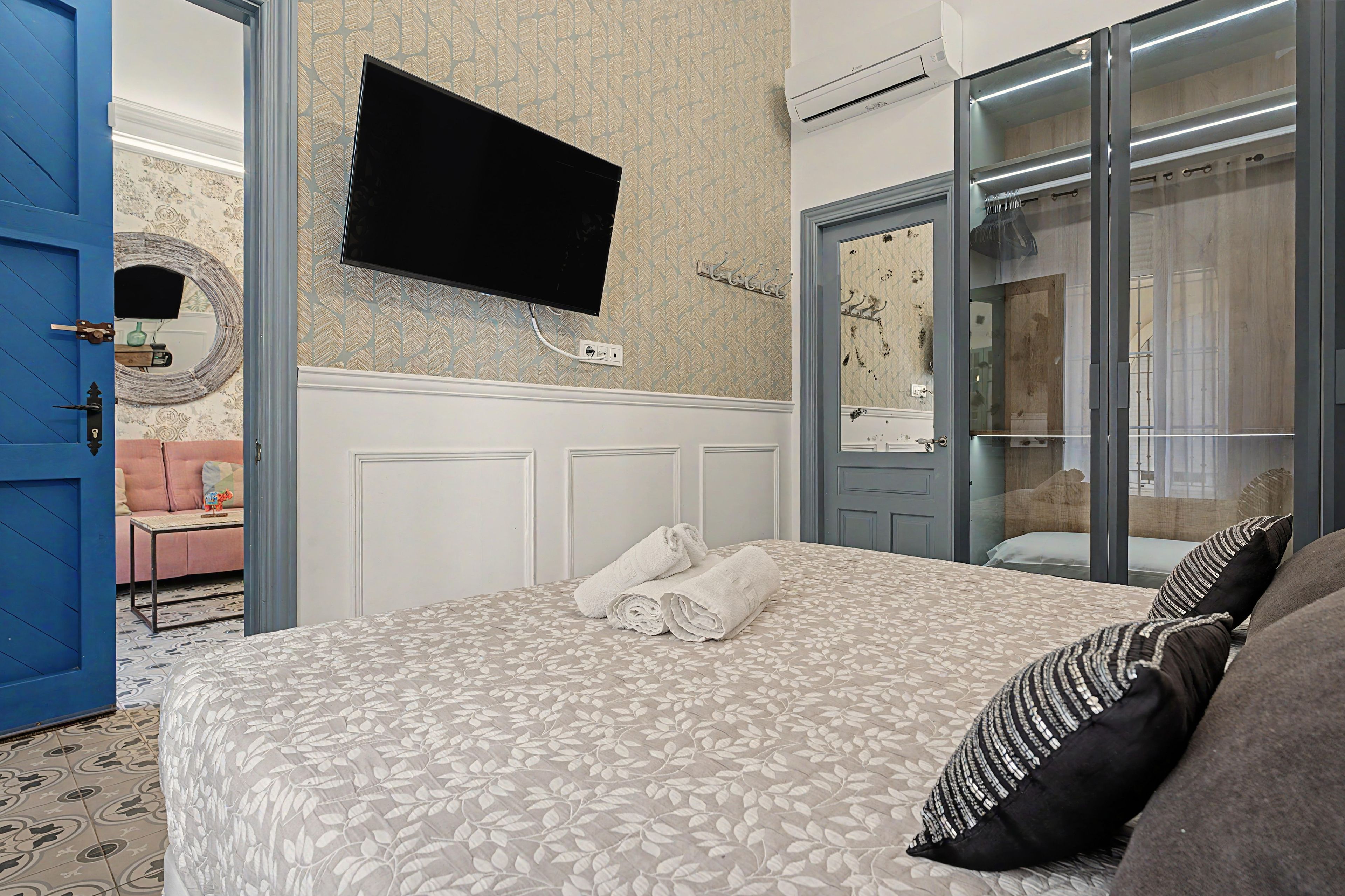 Quarto de hotel com cama, televisão de tela plana na parede, guarda-roupa com espelho e banheiro visível através de uma porta de vidro.