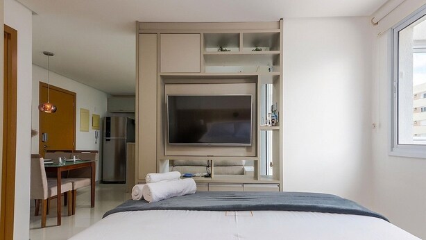 A hotel room with a bed, a TV, a desk with a chair, and a kitchenette area. - Apartment in a Condominium in Setor Bueno Lhd0403 (Goiânia)