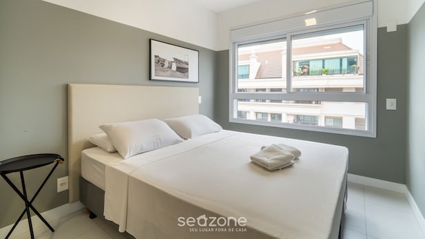 Quarto moderno com uma cama grande, uma mesa pequena, um quadro emoldurado e uma janela com vista para prédios.