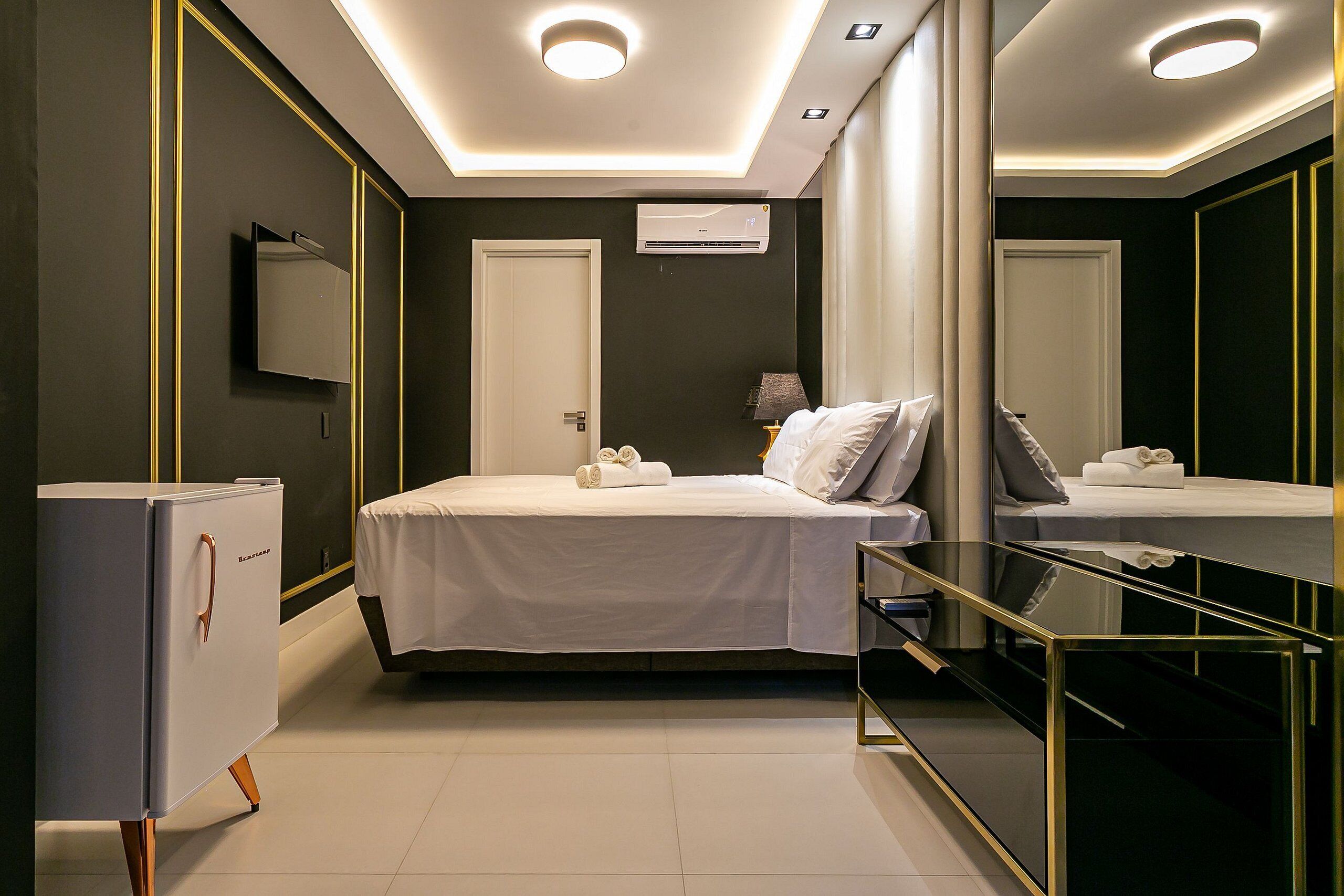 A modern hotel room with a bed, a minibar, a mirror, and a ceiling light.