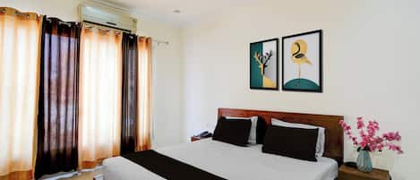 Une chambre d’hôtel avec un lit, des rideaux, un climatiseur fixé au mur, une table de chevet avec un téléphone et un vase avec des fleurs.