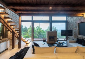 A modern living room with a stone fireplace, a sofa, a television, and a large window offering a view of greenery.