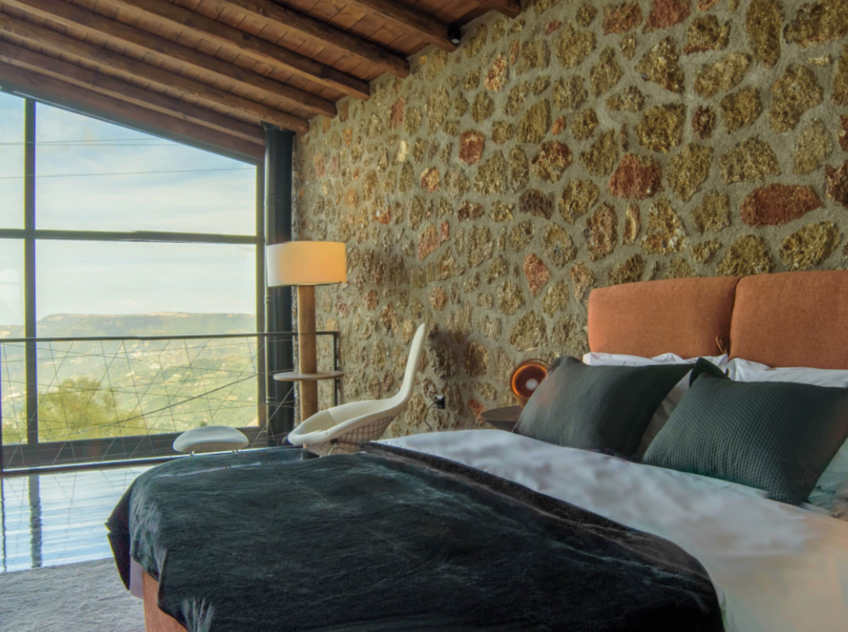 A bedroom with a stone wall, a bed with multiple pillows, a chair, and a large window offering a view.