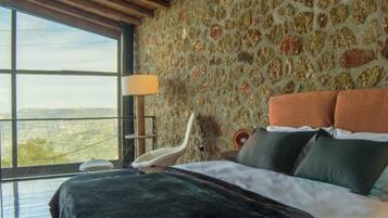 A bedroom with a stone wall, a bed with multiple pillows, a chair, and a large window offering a view.