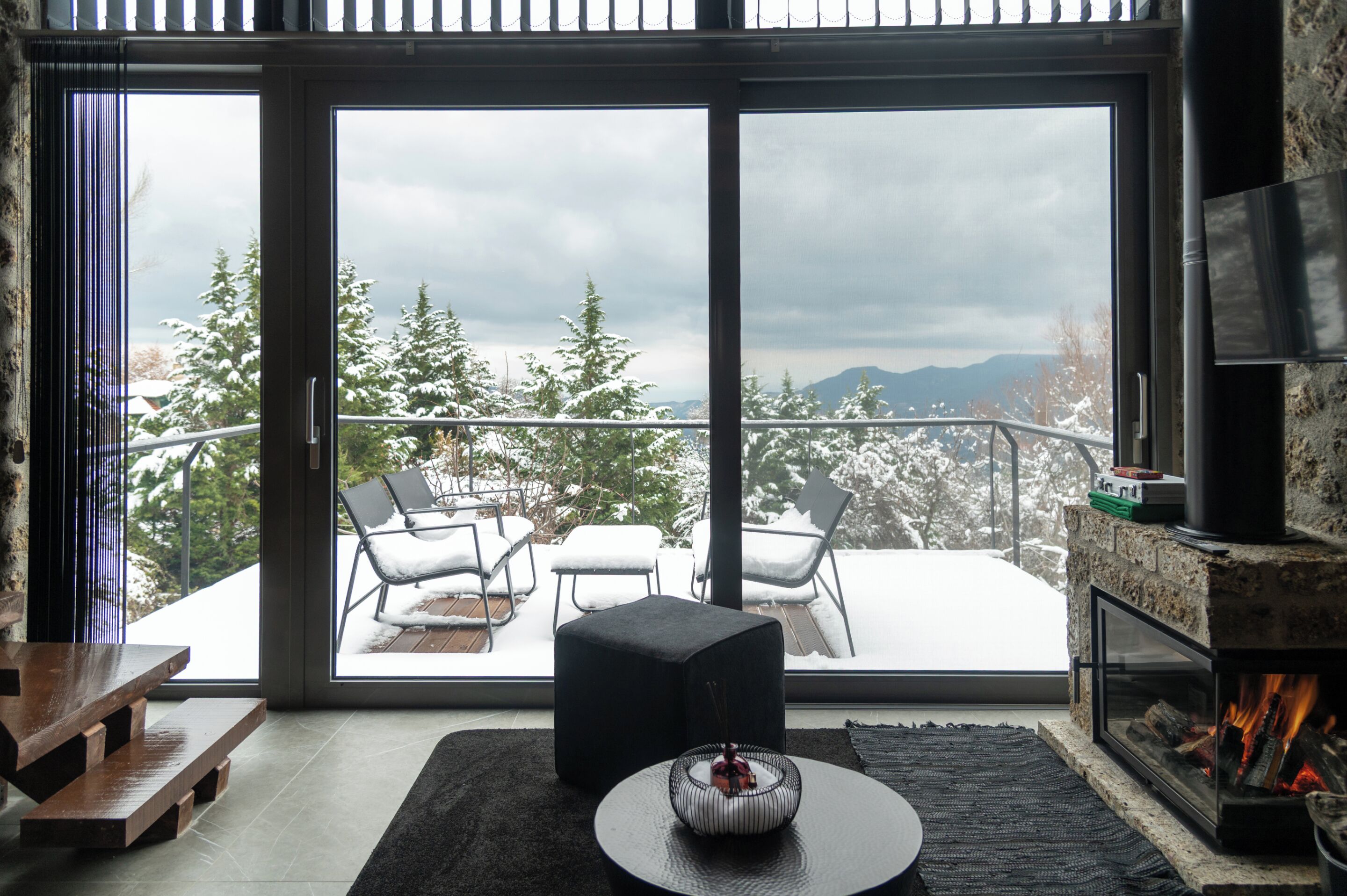 A modern living room with a fireplace, a large sliding glass door leading to a balcony with outdoor seating and snow-covered trees.