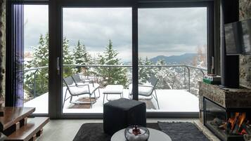 A modern living room with a fireplace, a large sliding glass door leading to a balcony with outdoor seating and snow-covered trees.