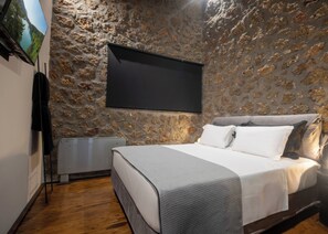 Quarto com parede de pedra, cama com roupa de cama branca, televisão de tela plana na parede e radiador.