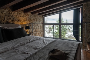 A bedroom with a large window, a bed, a bedside table with a lamp, and a stone wall.