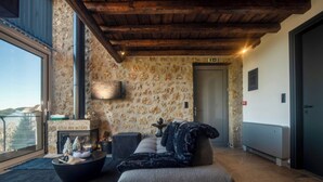 A cozy living area with a stone wall, a fireplace, a sofa, and a view through a large window. - Heartbeat Mountain Escapes (Kato Sinikia Trikalon)