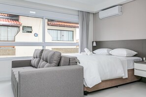 A modern hotel room with a bed, a grey sofa, and a window with a view of buildings. - Complete Studio 3 Minutes From the sea Jrr205 (Florianópolis)